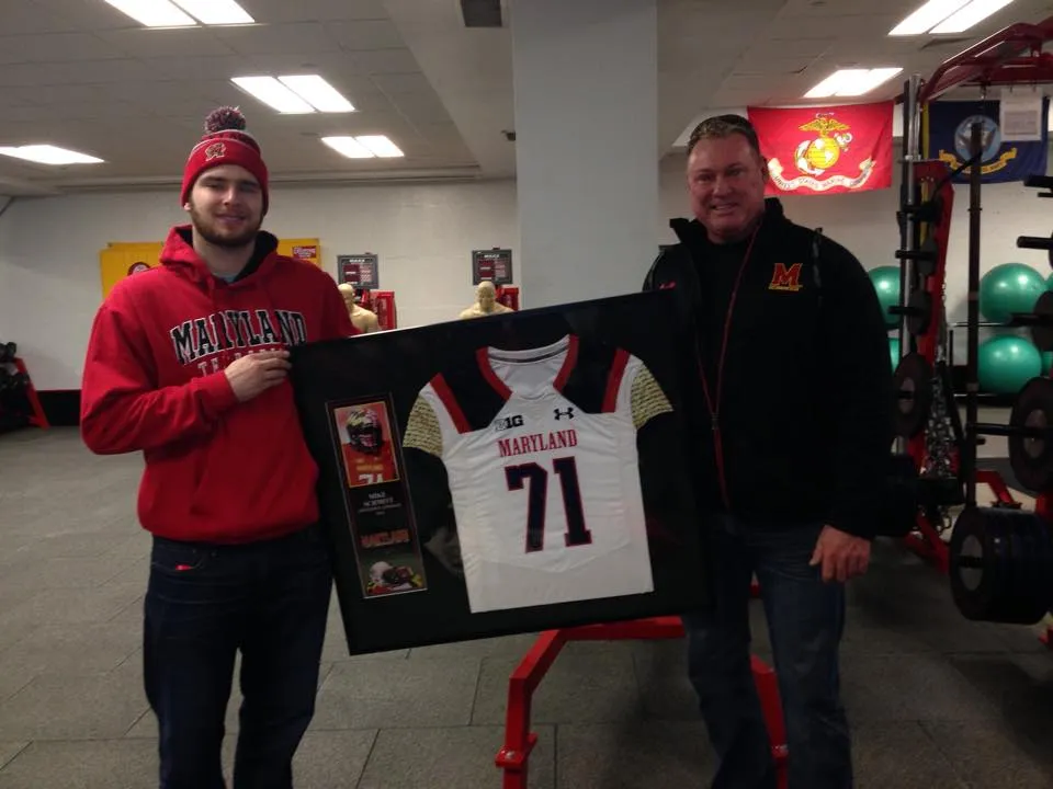 Two men in Maryland gear hold a framed Maryland football jersey inside a gym with weights and fitness equipment.